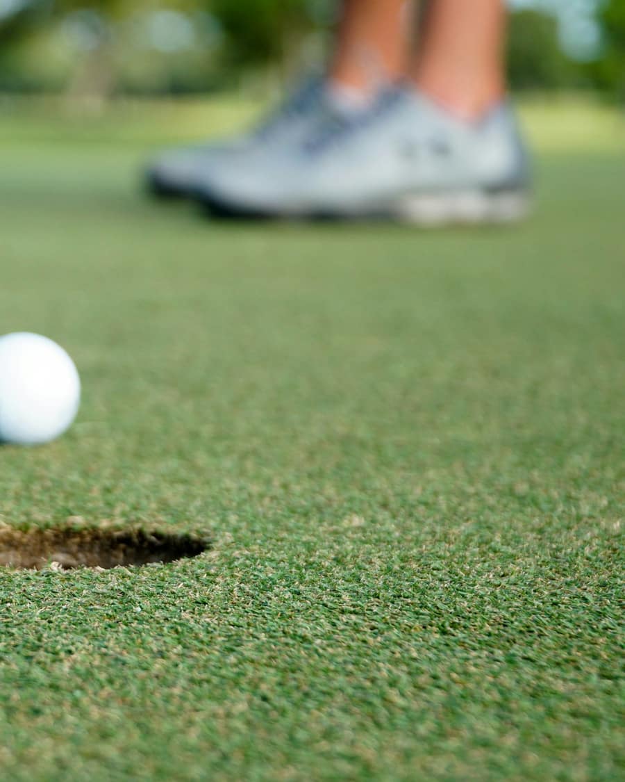A golf ball an inch from the hole on a short green, with a putter swinging in behind it